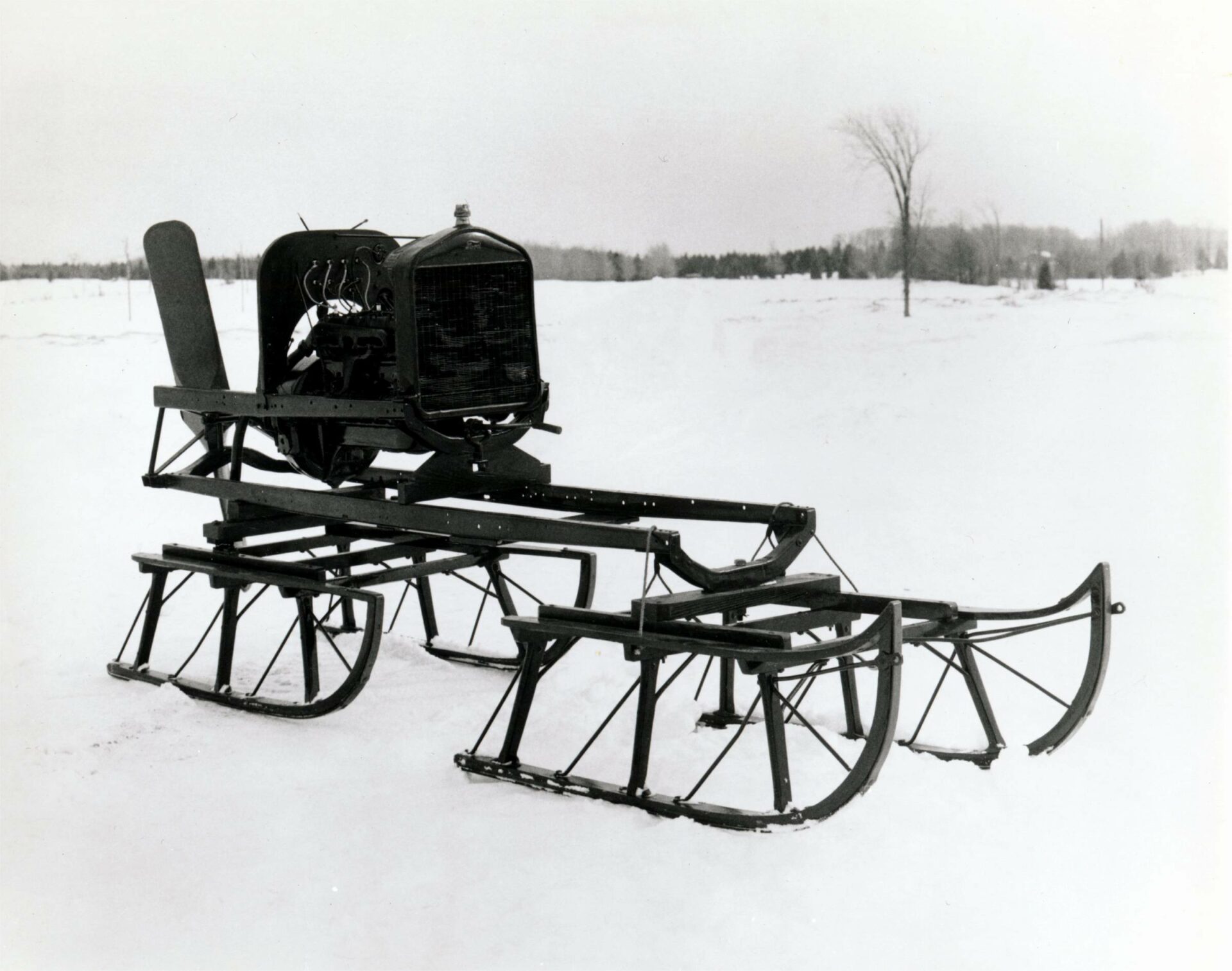 Photographie en noir et blanc du véhicule 1922 en hiver vu de profil. Le véhicule est muni d’une l’hélice à l’arrière, d’un moteur Ford Modèle T au centre et de quatre patins de traîneau fixés sur une armature de métal.