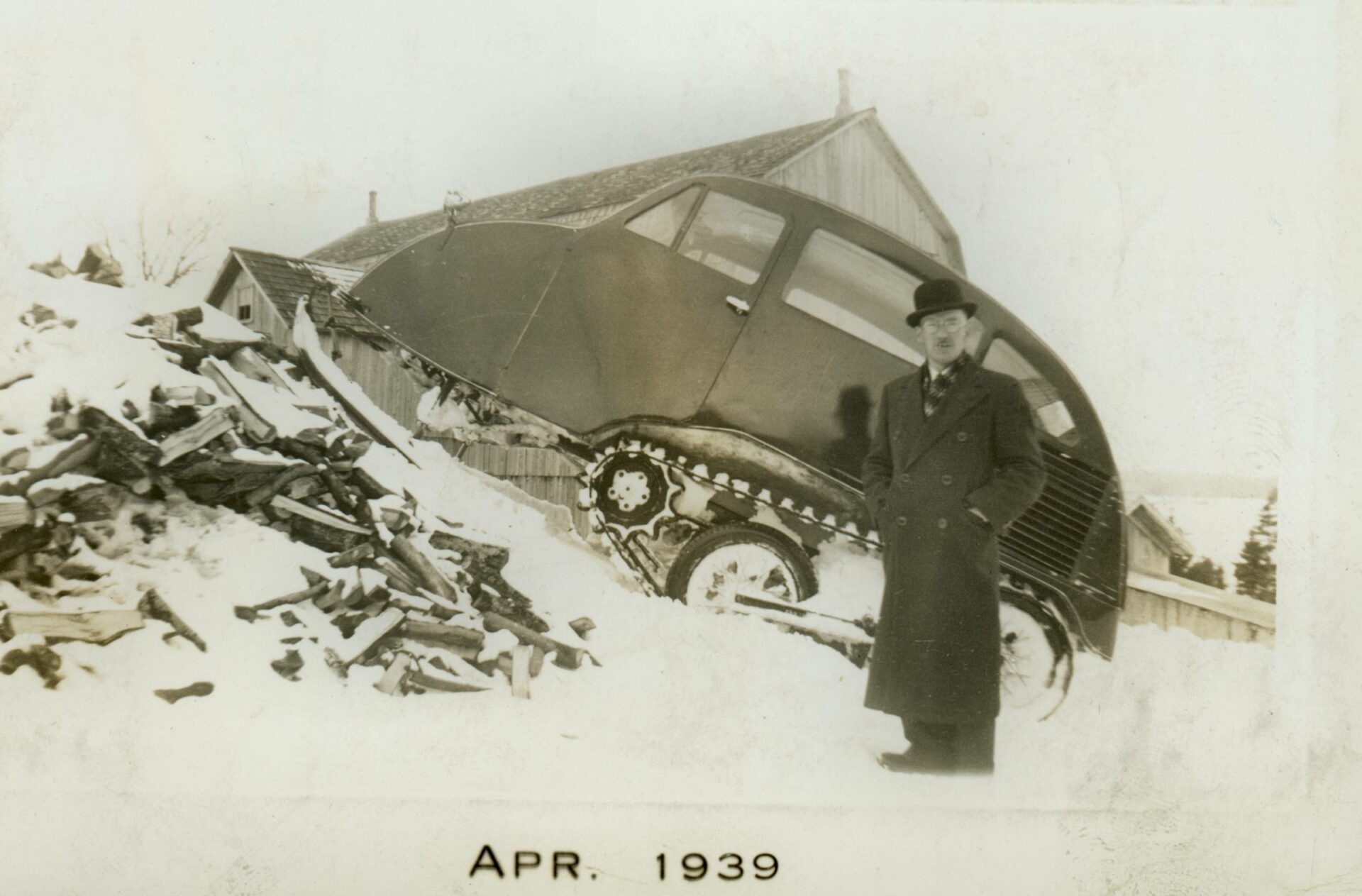 Photographie en noir et blanc de Joseph-Armand Bombardier devant une autoneige B7 en hiver. Vue de profil elle est montée sur un tas de bois enneigé. De forme arrondie, elle a quatre fenêtres, deux skis à l'avant et est munie du système de traction barbotin-chenille. L’inventeur porte un chapeau melon et un long manteau d’hiver.