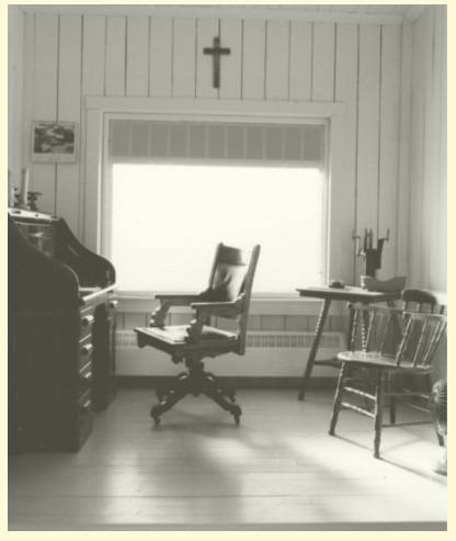 Photographie en noir et blanc du bureau de Joseph-Armand Bombardier dans le Garage Bombardier. À gauche il y a un secrétaire en bois et une chaise sur roulettes. Au fond, un mur en latte comprend une fenêtre, un cadre, un crucifix et un calorifère. À droite il y a une petite table avec des accessoires de bureau et une chaise avec accoudoirs.