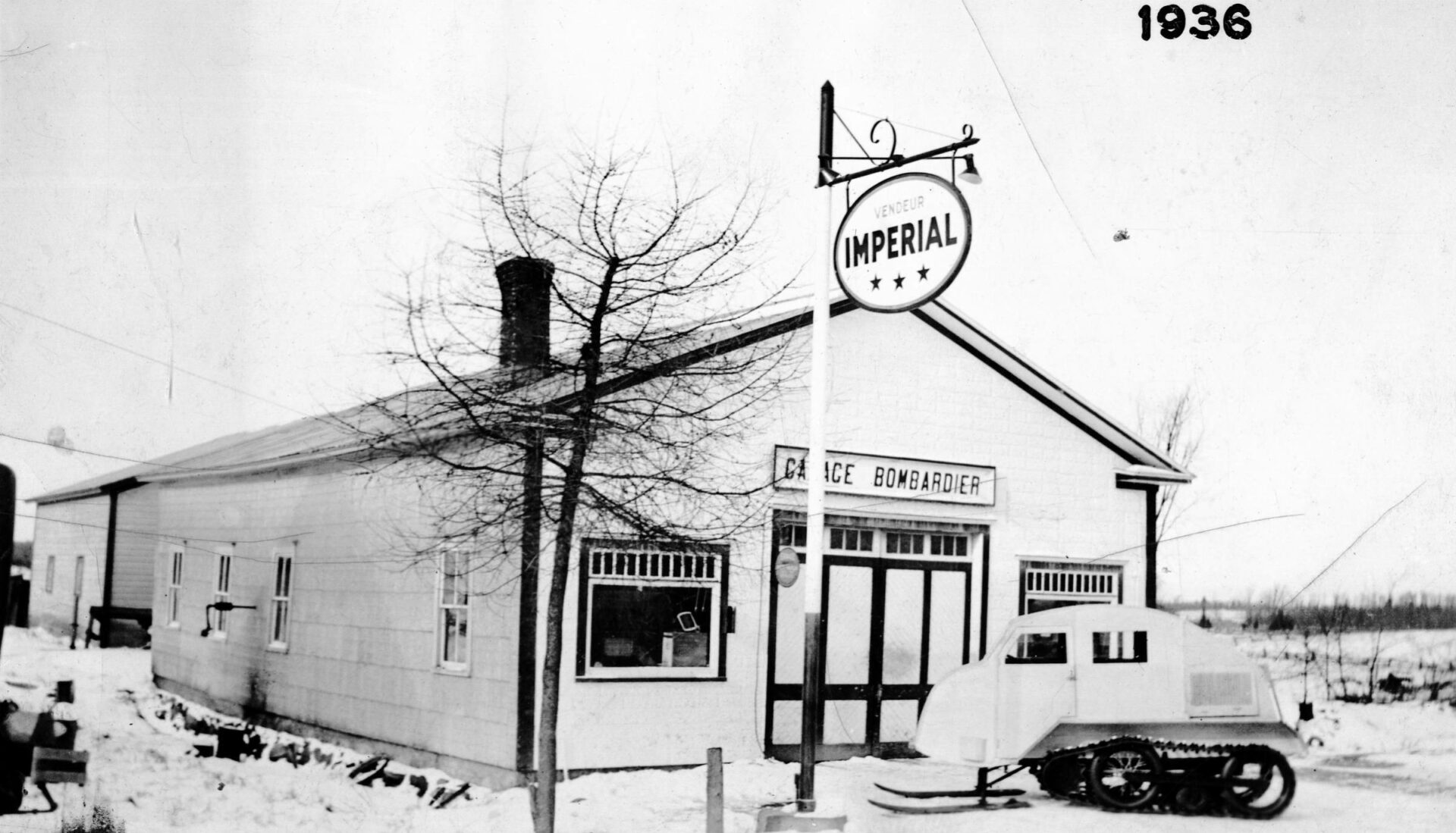 Photographie en noir et blanc du Garage Bombardier en hiver avec quatre fenêtres sur le côté gauche, deux fenêtres et une porte sur la façade. Une enseigne Vendeur Imperial figure aux côtés d’un prototype de l’autoneige B7, véhicule de forme arrondie avec des skis et un système de traction barbotin-chenille.