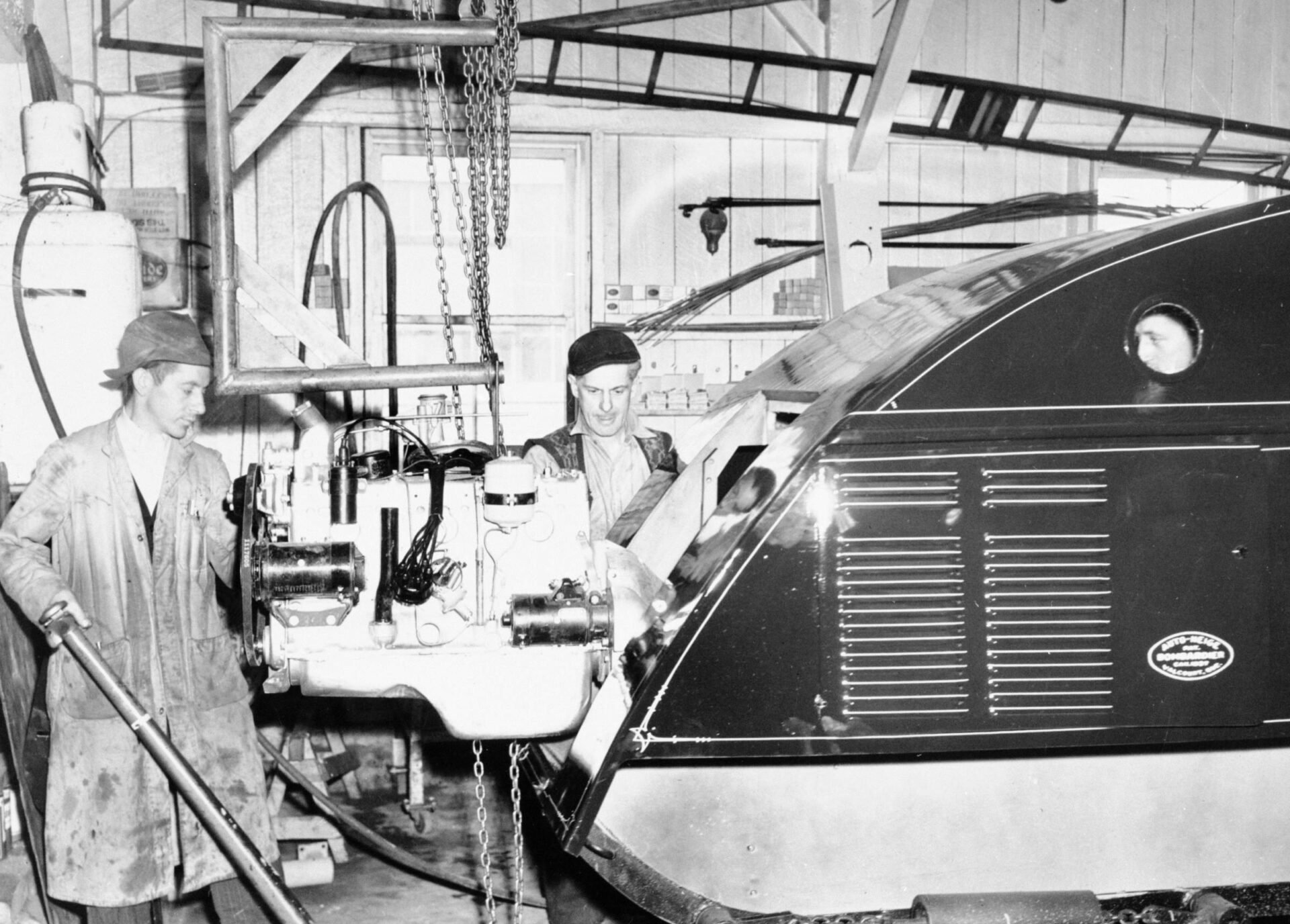 Photographie en noir et blanc de l’intérieur du Garage Bombardier. Aidés d’un support à moteur et d’un bloc de chaîne suspendu au plafond, trois hommes assemblent un moteur à l’arrière d’une autoneige B12. Ils portent des habits de garagiste. On voit le visage de l’un deux au travers le dernier hublot du véhicule.