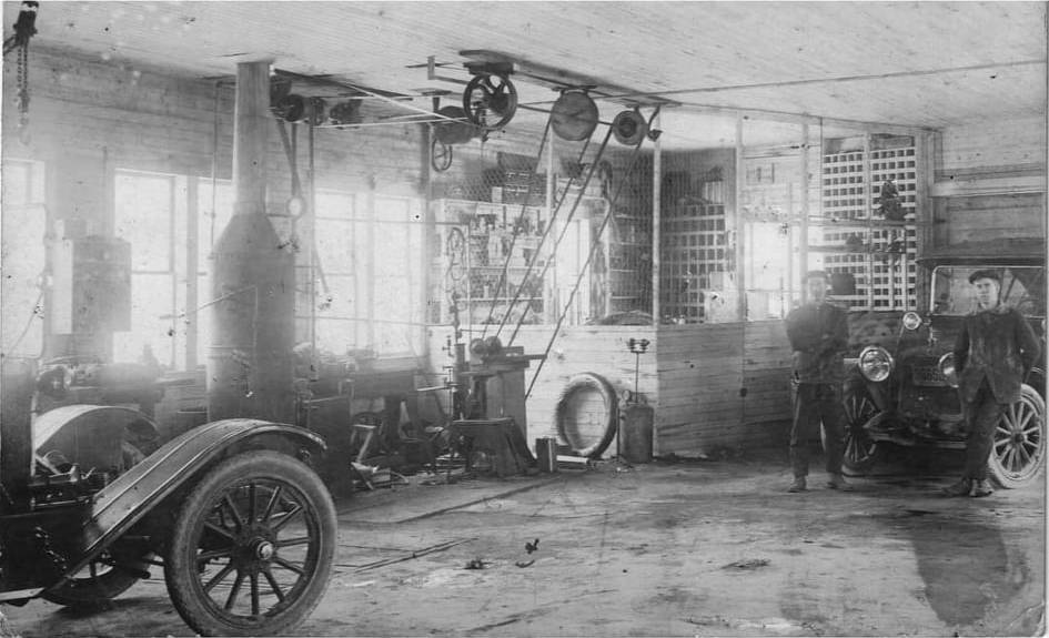 Black and white interior photograph of Garage Bombardier. On the right, two men dressed in 1920 typical attire with a coat and woolly cap, are posing in front of a Ford T automobile. On the left we can see a parts shop, a tire, garage tools, ceiling pulleys, a stove, and part of a car.