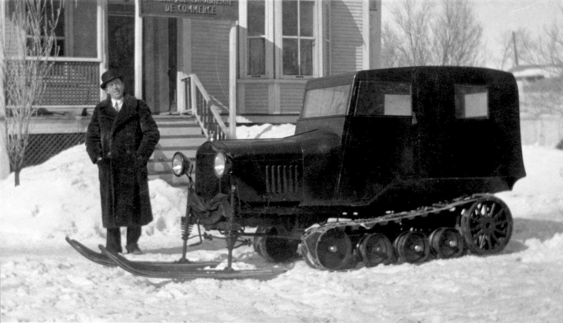 Photographie en noir et blanc du véhicule 1931 en hiver vu de profil et du notaire Antonio Grandpré de Valcourt devant la Banque Canadienne de commerce. Le véhicule Ford Modèle T est muni de deux skis à l’avant, d’une suspension composée de roues et de chenilles souples à l’arrière. Le notaire porte un manteau de fourrure et un chapeau melon.