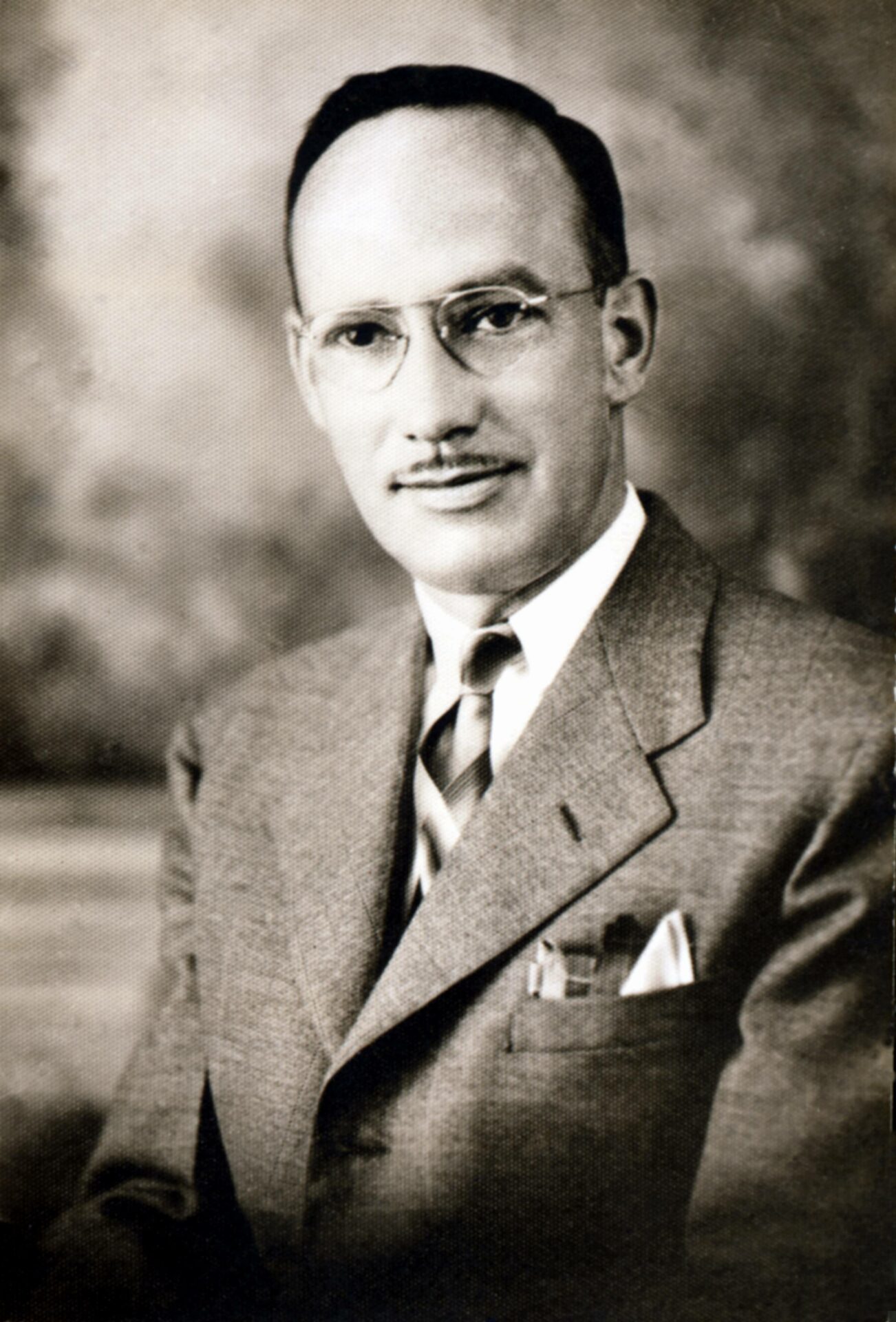 Photographie en noir et blanc, portrait de Joseph-Armand Bombardier. Il porte un veston, une chemise blanche et une cravate raillée ainsi qu’un mouchoir de poche. M. Bombardier porte des lunettes et il a une moustache.