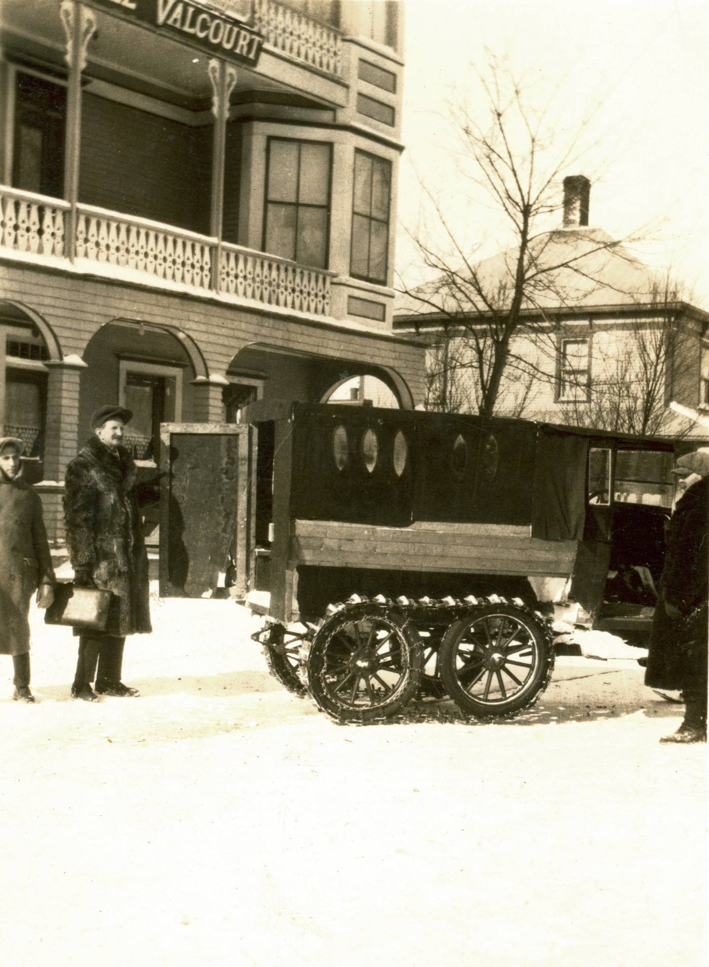 Photographie en noir et blanc de trois hommes devant l’hôtel de Valcourt en hiver aux côtés d’un véhicule muni de cinq hublots et d’un système de chenille de métal sur des roues à rayons. Ils portent des manteaux de fourrure. Au centre, le Docteur Phydime Langlois tient une mallette médicale à la main droite.