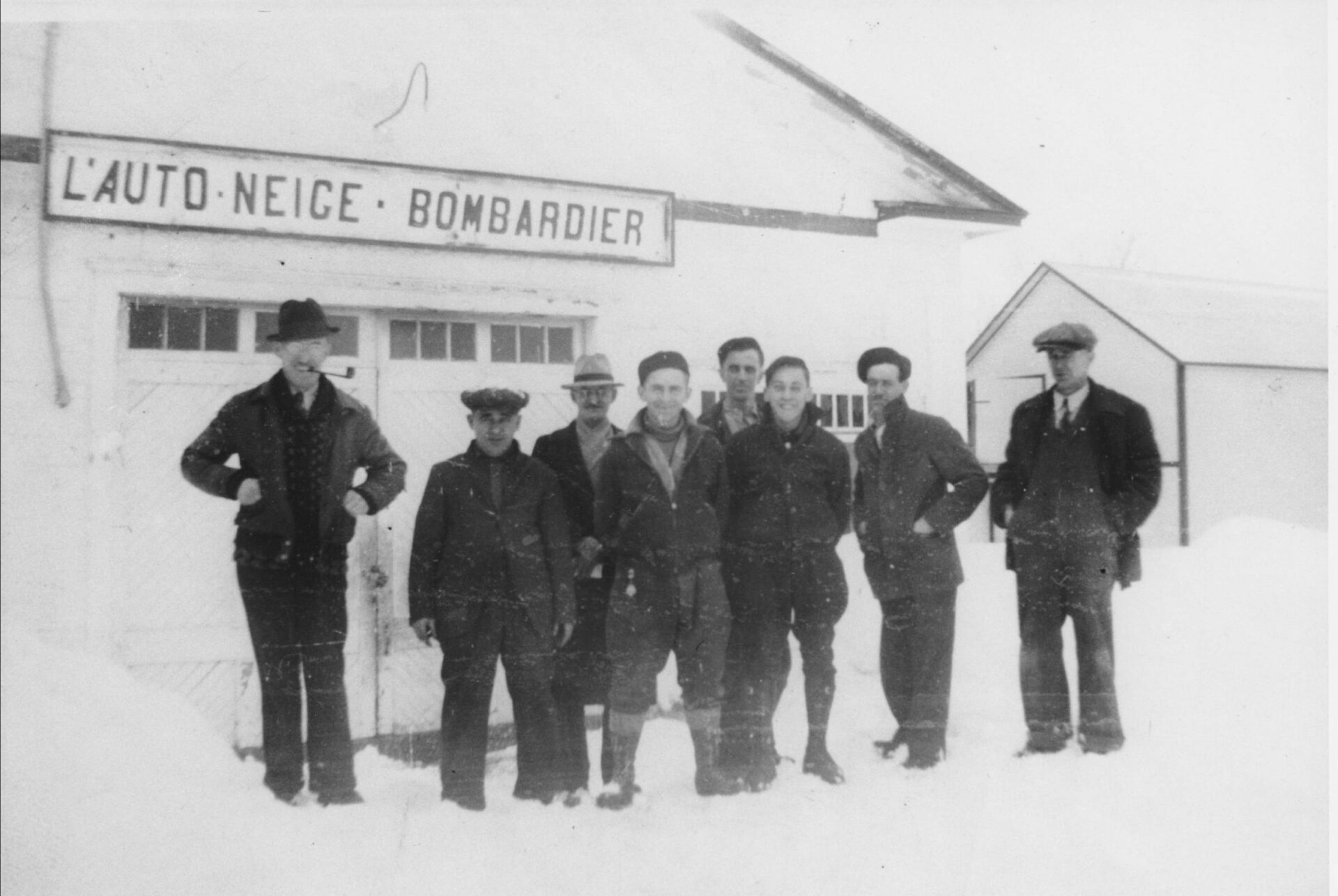 Photographie en noir et blanc des premiers employés de Joseph-Armand Bombardier en hiver devant L’Auto-Neige Bombardier. Ils sont habillés pour le froid : casquettes, chapeaux et manteaux typiques aux années 1930. Joseph-Armand Bombardier la pipe à la bouche est souriant et porte chemise blanche et une cravate.