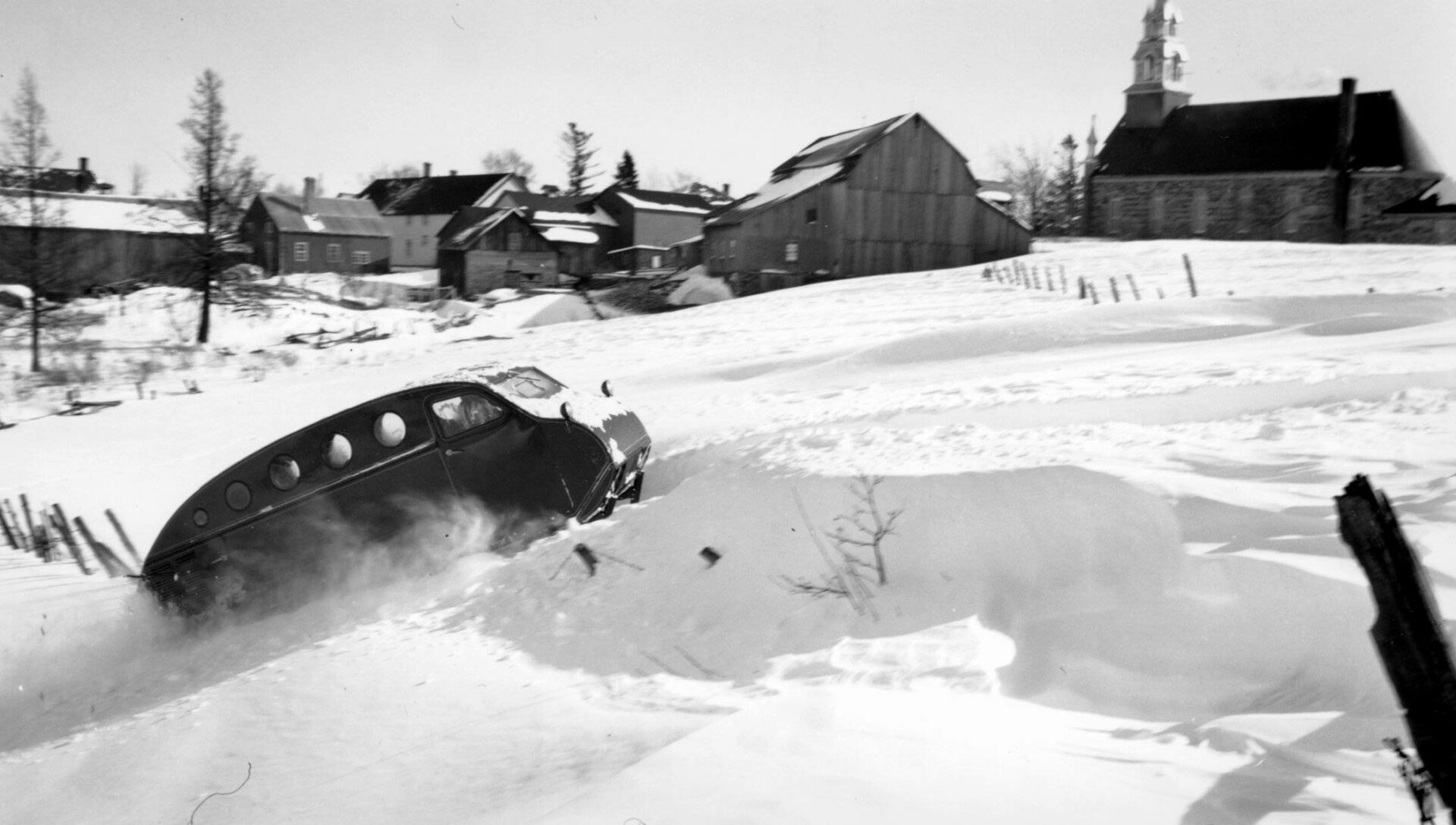 Photographie en noir et blanc d’une autoneige B12 circulant sur un champ enneigé de Valcourt. Le véhicule a la forme d’un autobus, cinq hublots sur le côté et des skis à l’avant. En arrière-plan, il y a des maisons, une grange et l’église du village.