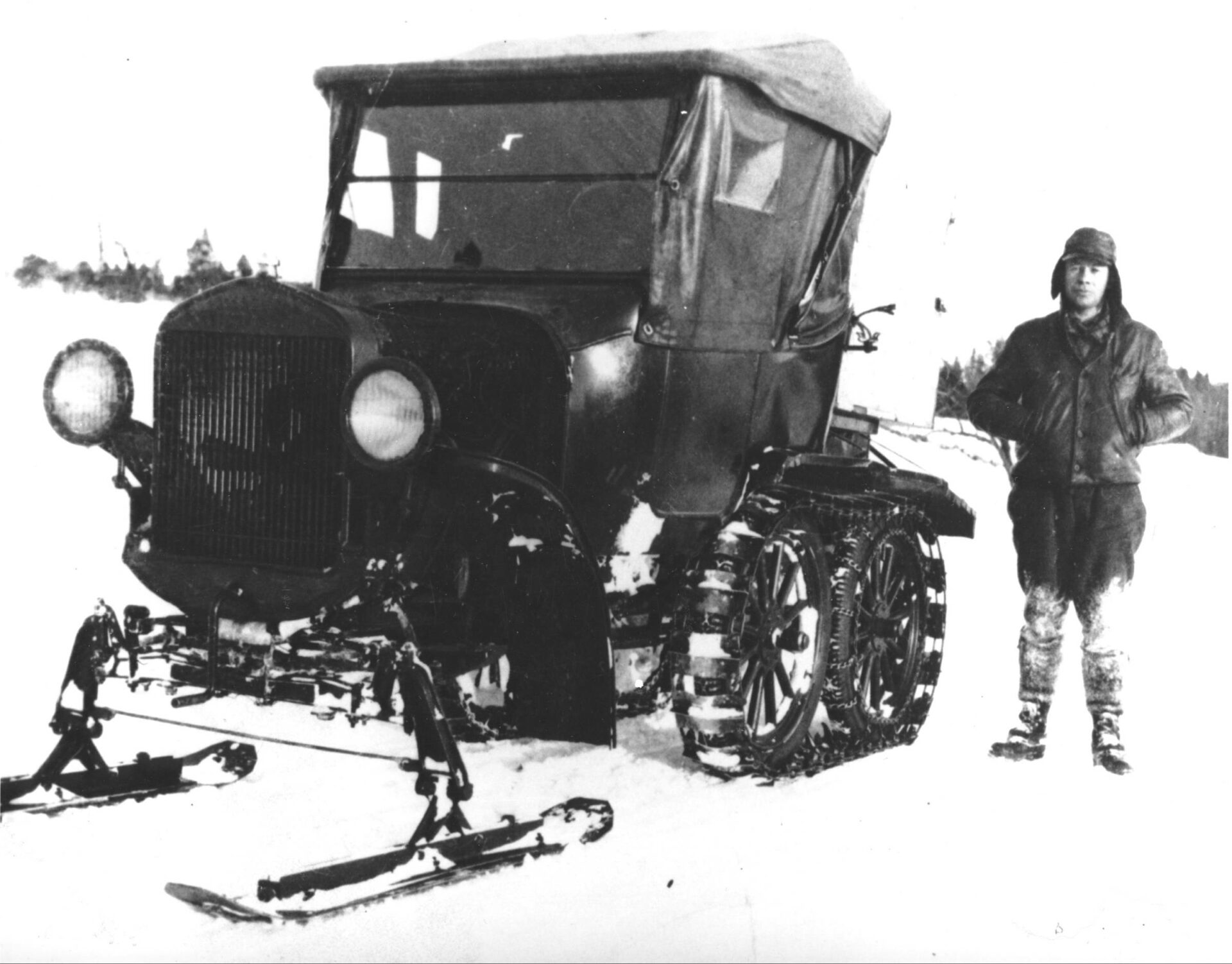 Photographie en noir et blanc de Valmore Labrecque aux côtés du véhicule 1929 développé par Joseph-Armand Bombardier. Le véhicule est muni de skis à l’avant et d’un système de chenilles de métal à l’arrière adapté à une voiture Ford Modèle T. M. Labrecque porte un chapeau de type aviateur et un habit de neige.