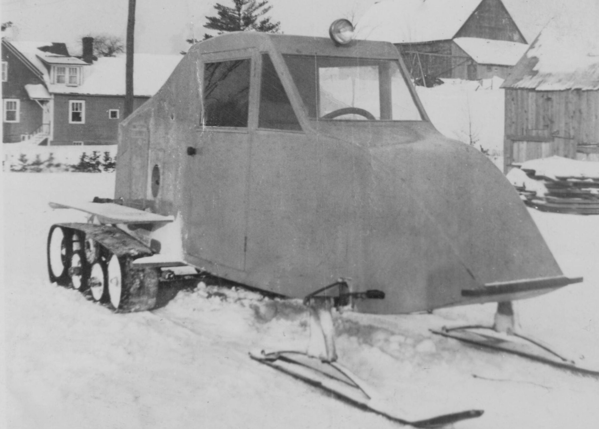 Photographie en noir et blanc du véhicule 1934 en hiver vu de profil. Le véhicule de forme aérodynamique est muni de deux skis à l’avant et de chenilles doubles souples entraînées par des roues à l’arrière. Le véhicule est muni d’une lumière en haut au centre. En arrière-plan, il y a une maison et des bâtiments de ferme.