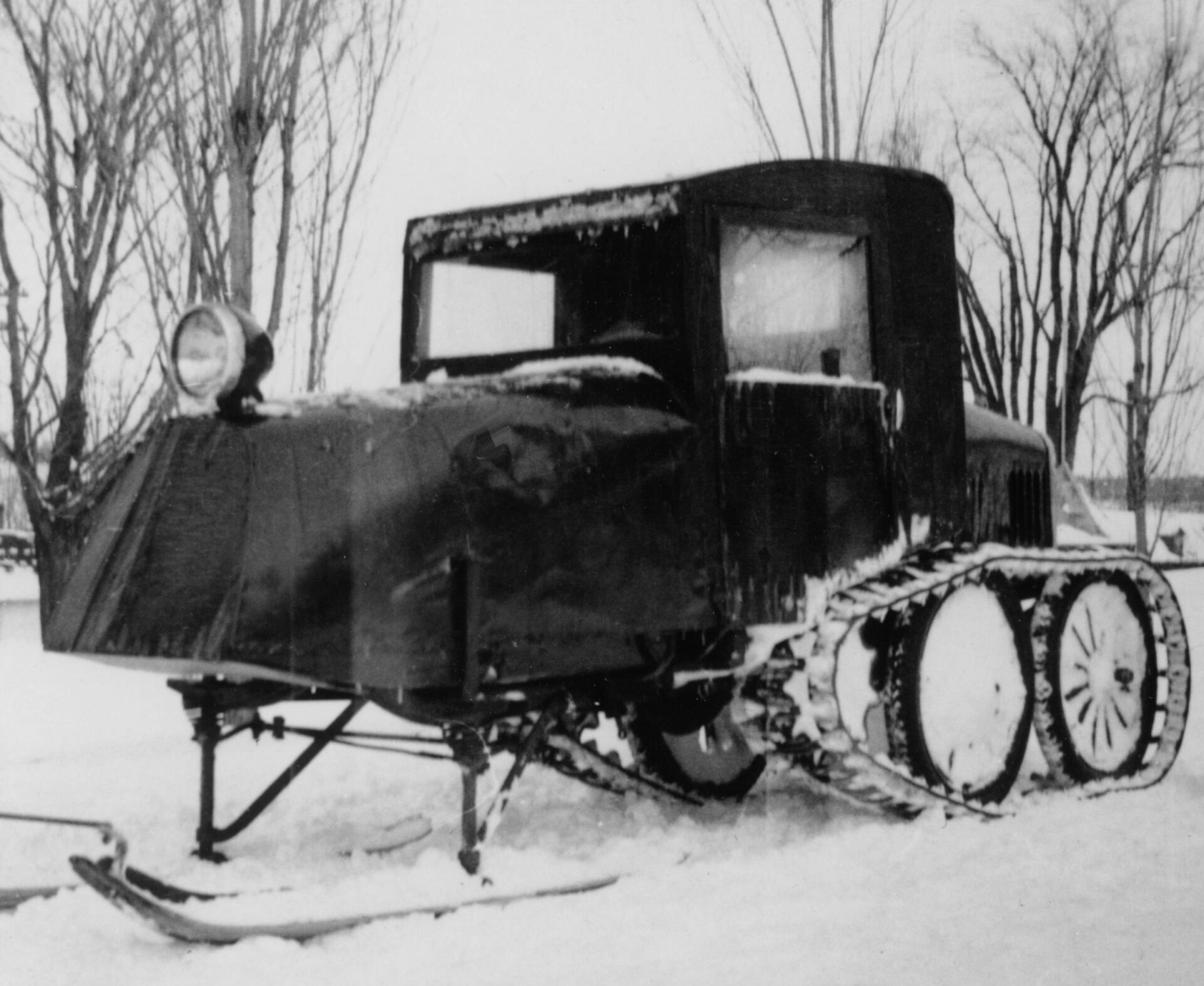 Photographie en noir et blanc du véhicule 1935 en hiver vu de profil. Le véhicule Ford modèle T est muni de deux skis à l’avant et du système de traction barbotin-chenille enneigé à l’arrière composé de roues d’engrenage dentées entraînant des chenilles souples de caoutchouc.