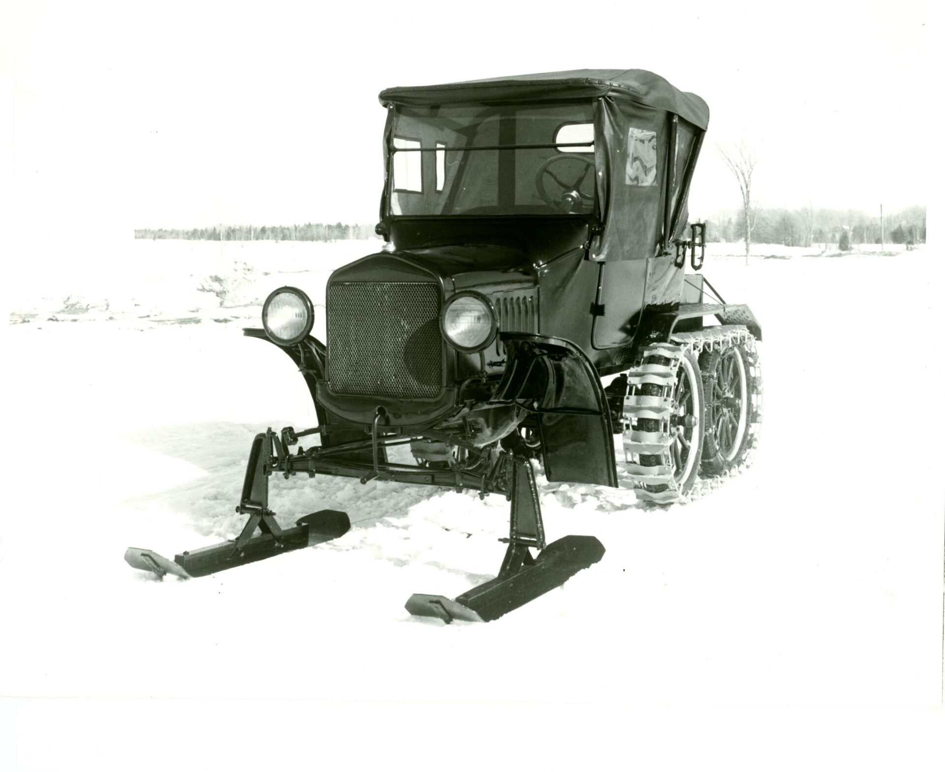Photographie en noir et blanc du véhicule 1929 en hiver vu de profil. Le véhicule Ford Modèle T est muni de deux skis à l'avant et d’un système de chenilles de métal constitué de traverses d’acier reliées entre elles par des mailles de chaîne installée sur une paire de roues à rayons l’arrière.