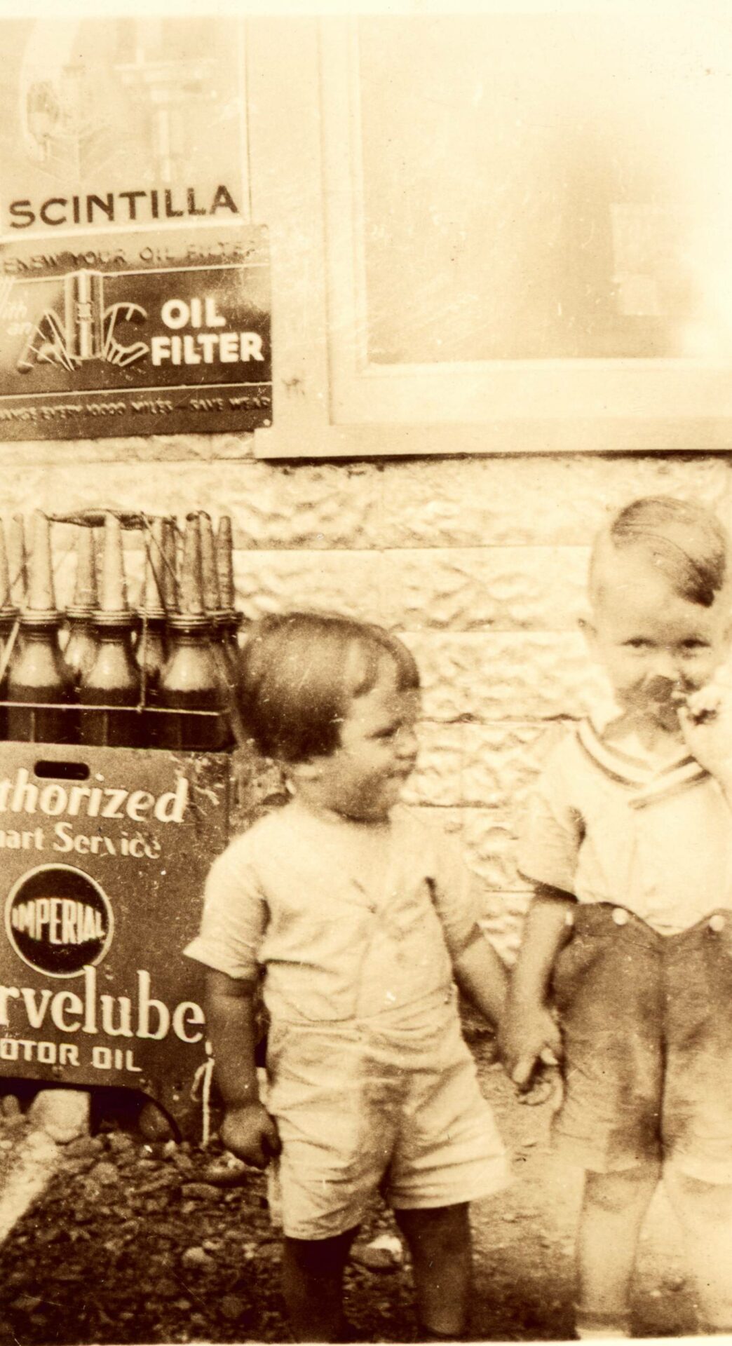 Photographie en sépia de Yvon et Germain Bombardier. Les enfants se tiennent la main et posent devant le Garage Bombardier. On voit des enseignes de filtreur et de moteur à l'huile. Ils portent des chandails et des culottes courtes.
