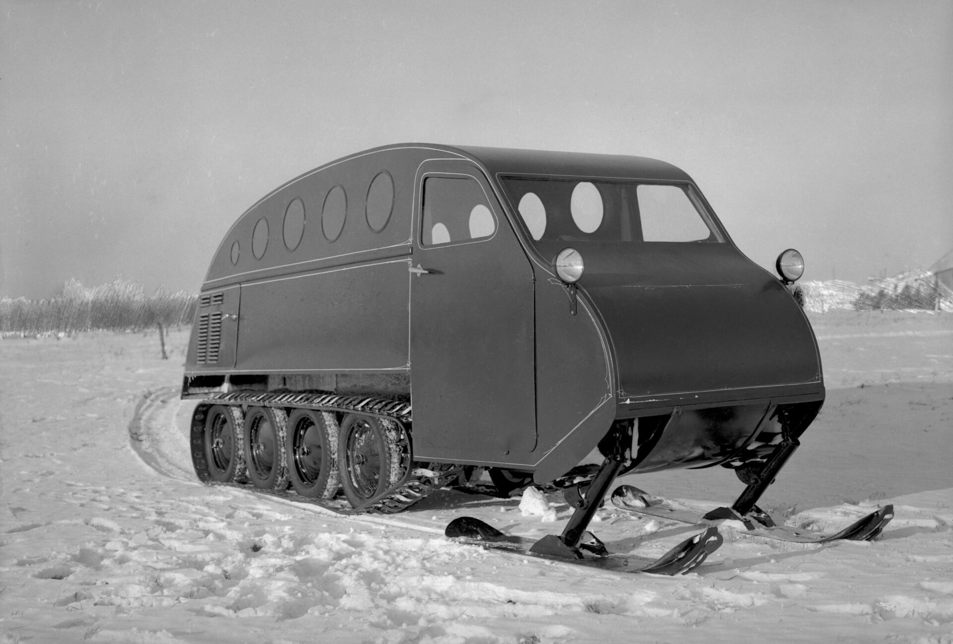 Photographie en noir et blanc d’une autoneige B12 en hiver vue de profil. Le véhicule à la forme d’un autobus avec cinq hublots de chaque côté, trois fenêtres, deux skis et le système de traction barbotin-chenille avec quatre roues pleines en acier.