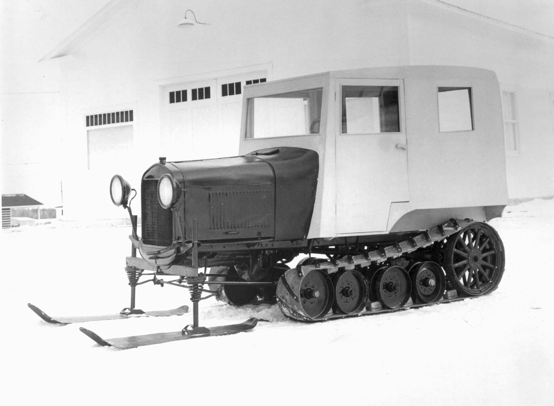 Photographie en noir et blanc d’une reconstitution du véhicule chenillé 1931 développé par Joseph-Armand Bombardier. Ce véhicule en question a été fabriqué par Alphonse-Raymond, Gérard et Léopold Bombardier, frères de Joseph-Armand Bombardier, vers 1968 dans le cadre du projet de la création du Musée J. Armand Bombardier.  On remarque sur la photo que le véhicule semble avoir deux couleurs, soit le noir pour la carrosserie et le rouge pour la cabine de bois ajoutée. Derrière le véhicule, on peut apercevoir le Garage Bombardier, en blanc. La photo a été prise à Valcourt sur le terrain de l’actuel Musée de l’ingéniosité J. Armand Bombardier. Le garage qui était originalement sur la rue Saint-Joseph y a été déménagé en 1968 pour la création du Musée.